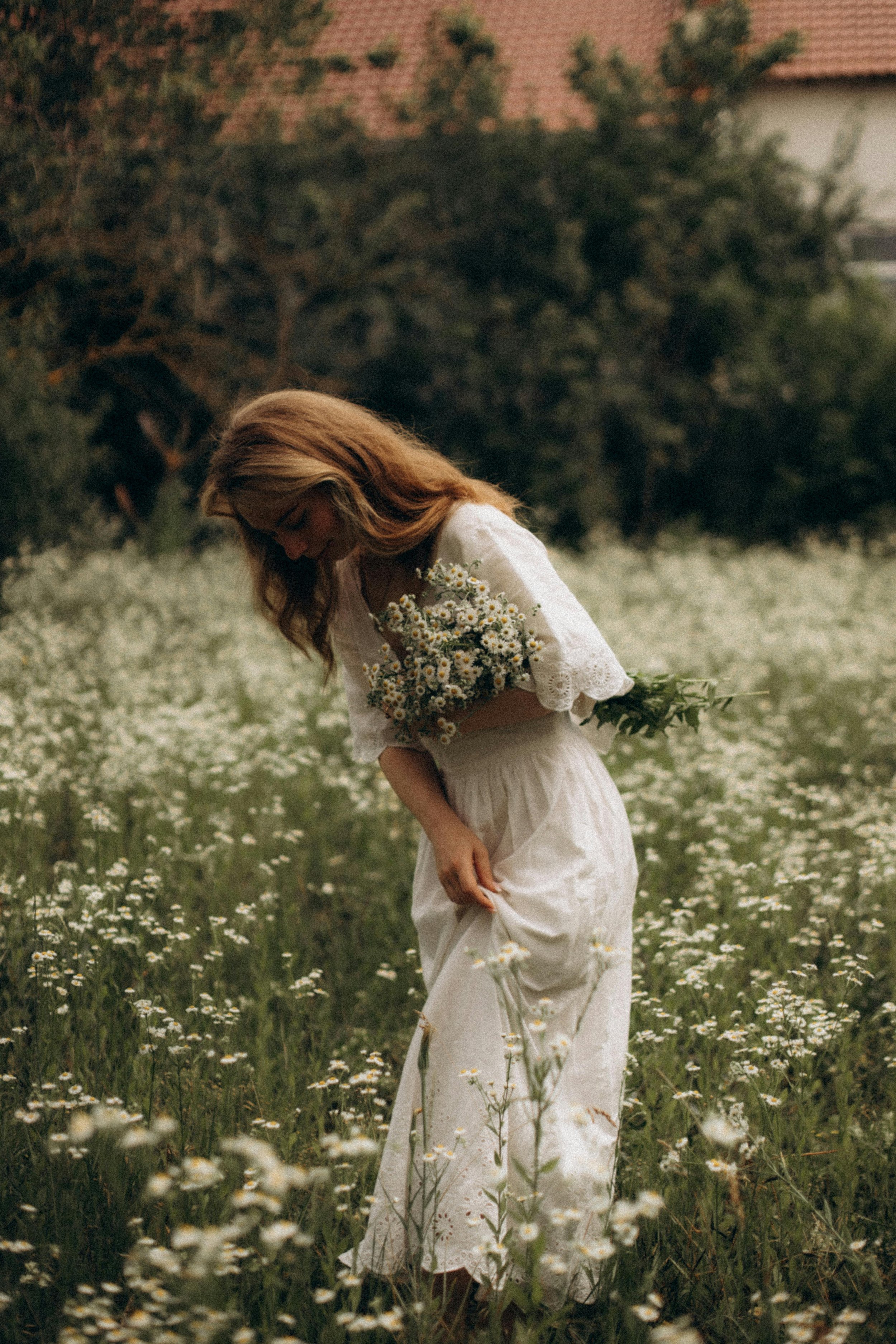 A woman in a white dress holding daisies, standing in a field of daisies with trees and a house in the background.