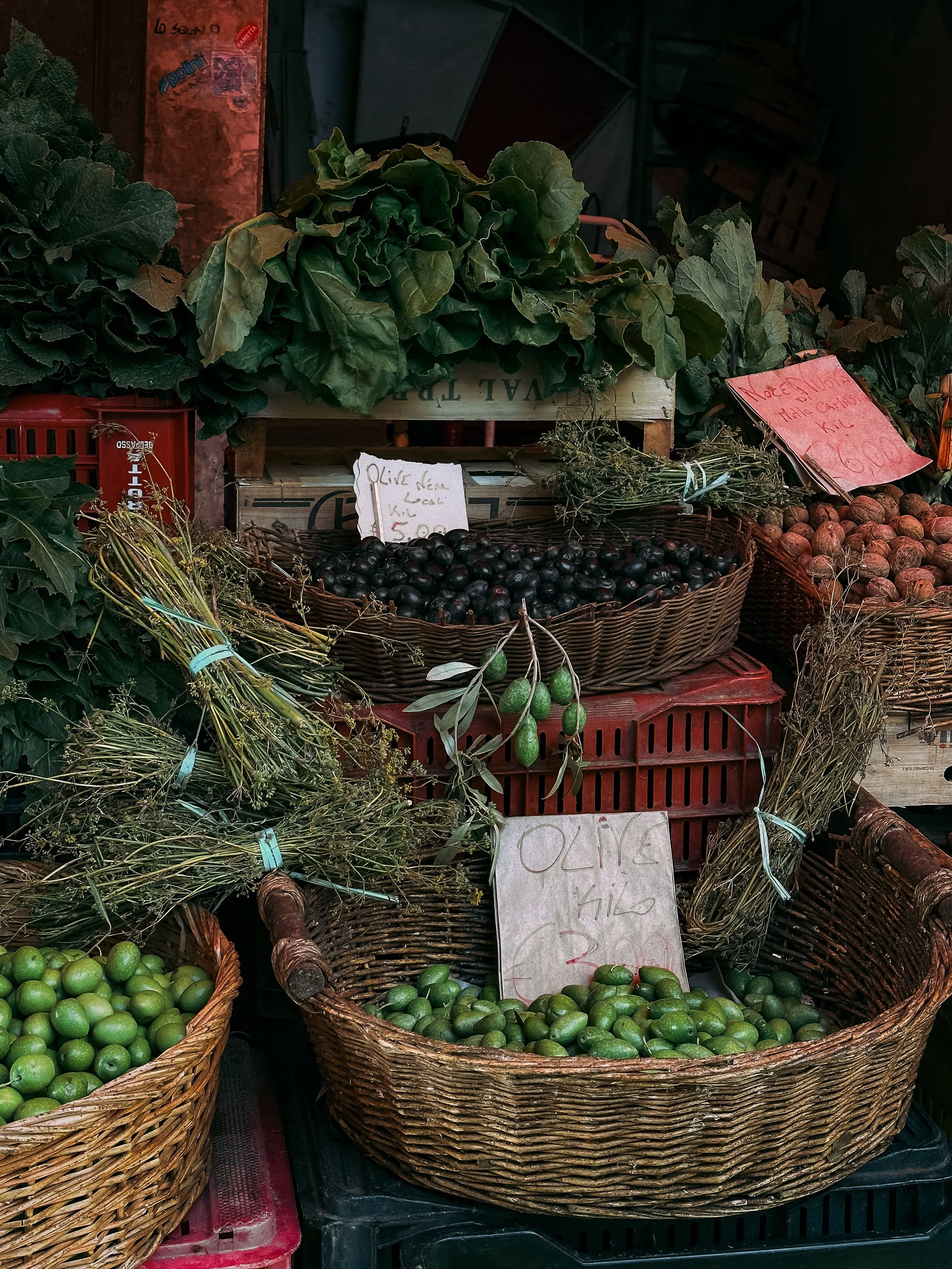 Baskets of limes, a basket of black olives, and other produce displayed at an outdoor market, with handwritten price signs.