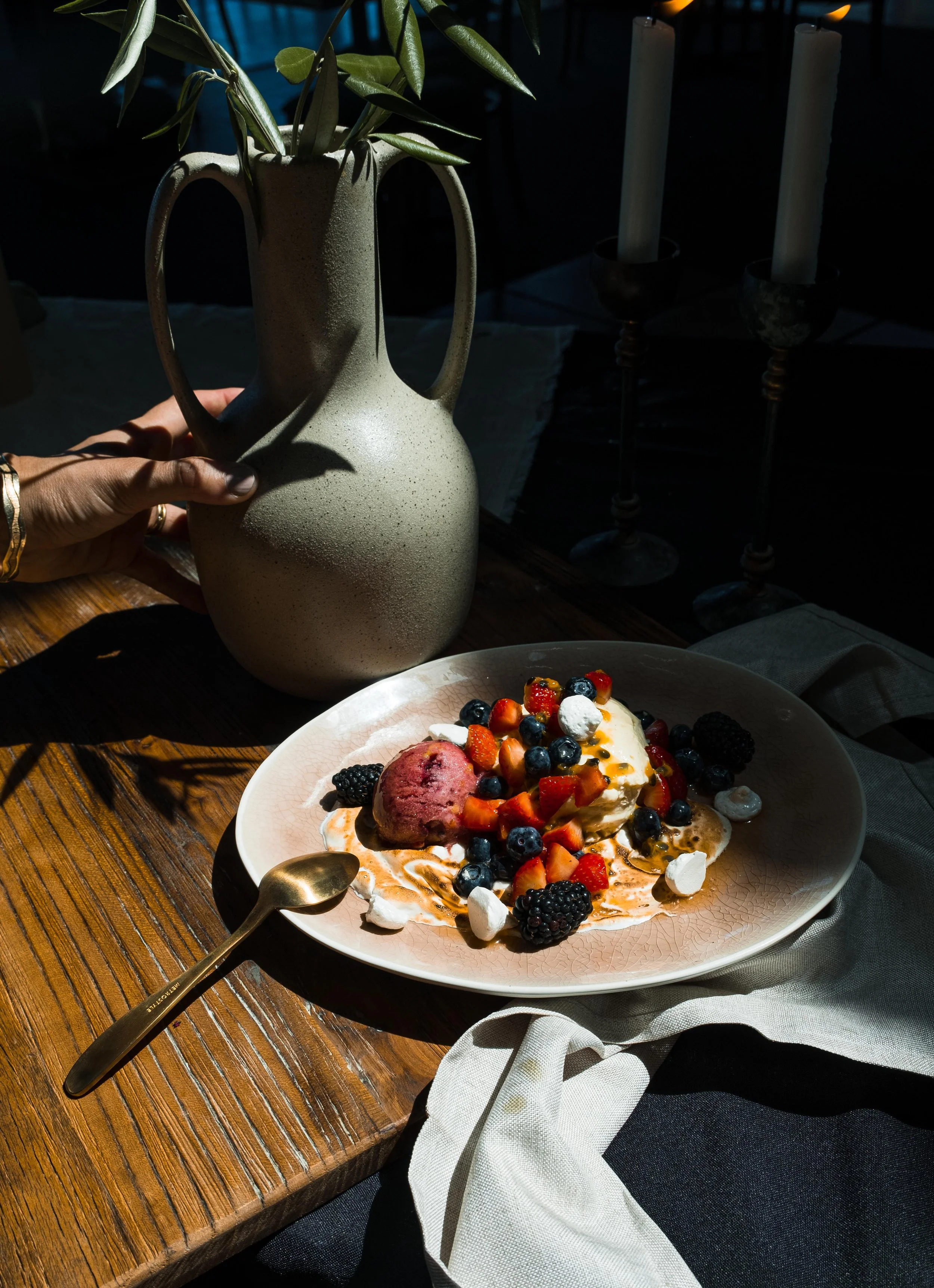 A beige ceramic pitcher with green leaves inside, being held by a hand with a gold bracelet, set on a wooden table. In the foreground, a plate of dessert topped with mixed berries, a dollop of cream, and syrup. A gold spoon rests on the table next to