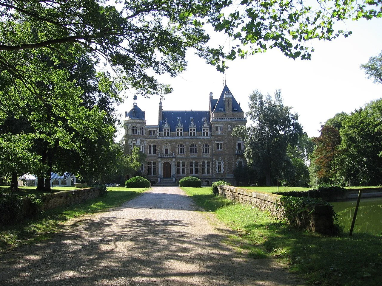 A large stone castle with turrets and towers, situated amidst lush green trees and grass, with a gravel pathway leading to its entrance, under a bright clear sky.