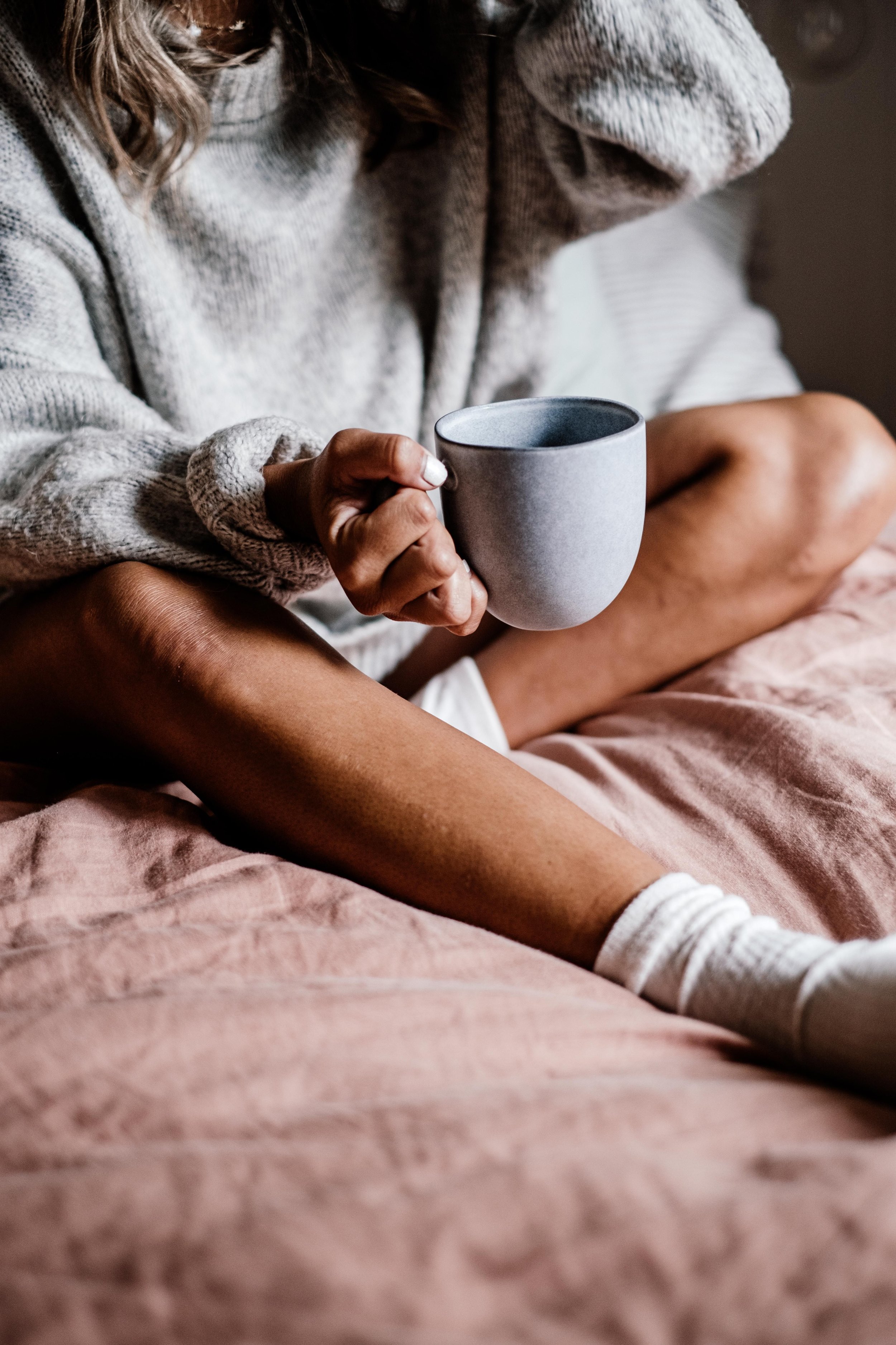 A person sitting cross-legged on a bed, holding a grey ceramic mug in their right hand, wearing a grey sweater, white socks, and a pink blanket covering their legs.