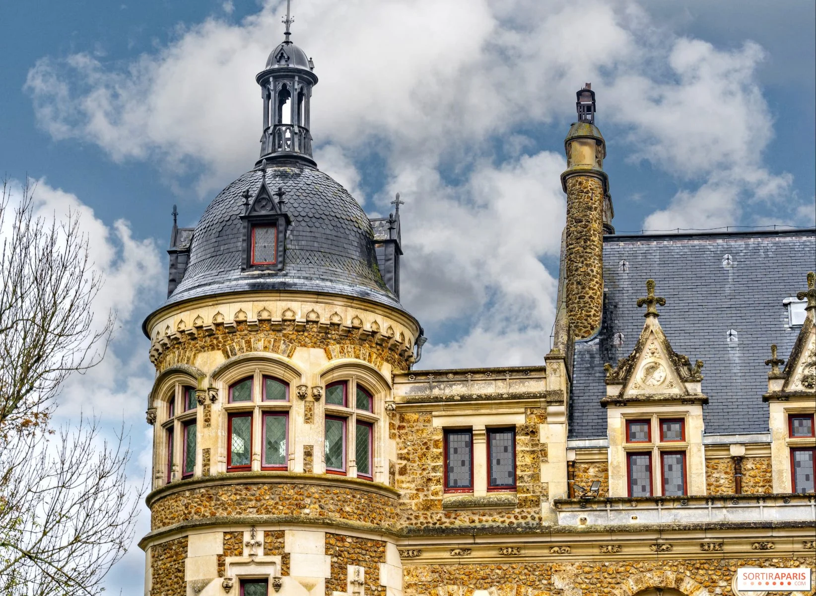 A close-up view of a historic castle or mansion with a turret and spire, with cloudy sky in the background and leafless trees in the foreground.