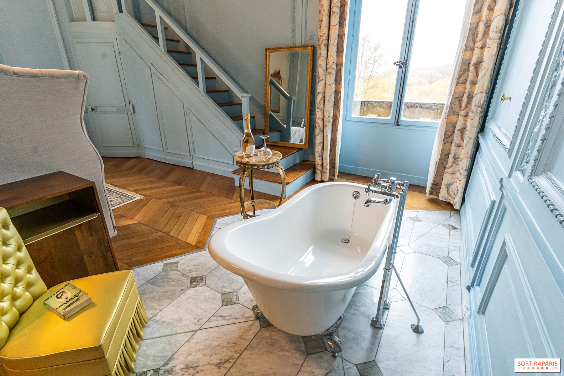 A vintage-style bathroom featuring a white clawfoot bathtub, a small ornate gold side table with a bottle and glasses, a large window with floral curtains, a staircase with white paneling, a mirror, a yellow bench with a book, and crown molding detai