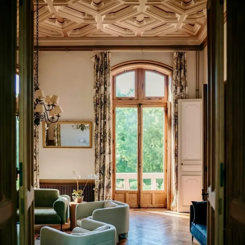 A bright living room with wooden framed French doors leading to a balcony and lush green trees outside, a patterned curtain on each side, a large mirror on the wall, a vintage chandelier, and seating with a couple of armchairs and a sofa.