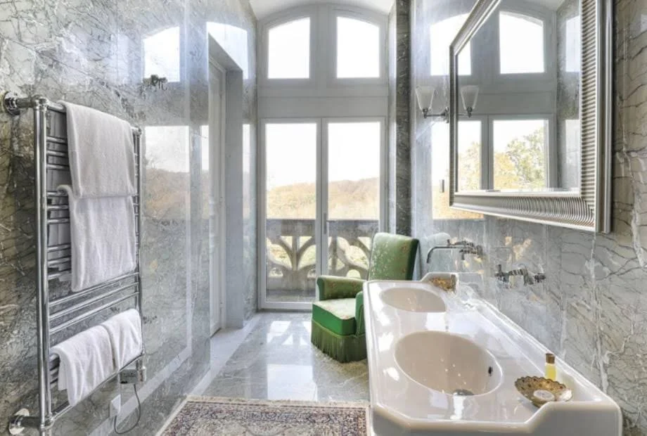 Bright bathroom with marble walls, a large window with a balcony view, a green armchair, a double sink countertop, and a large mirror.