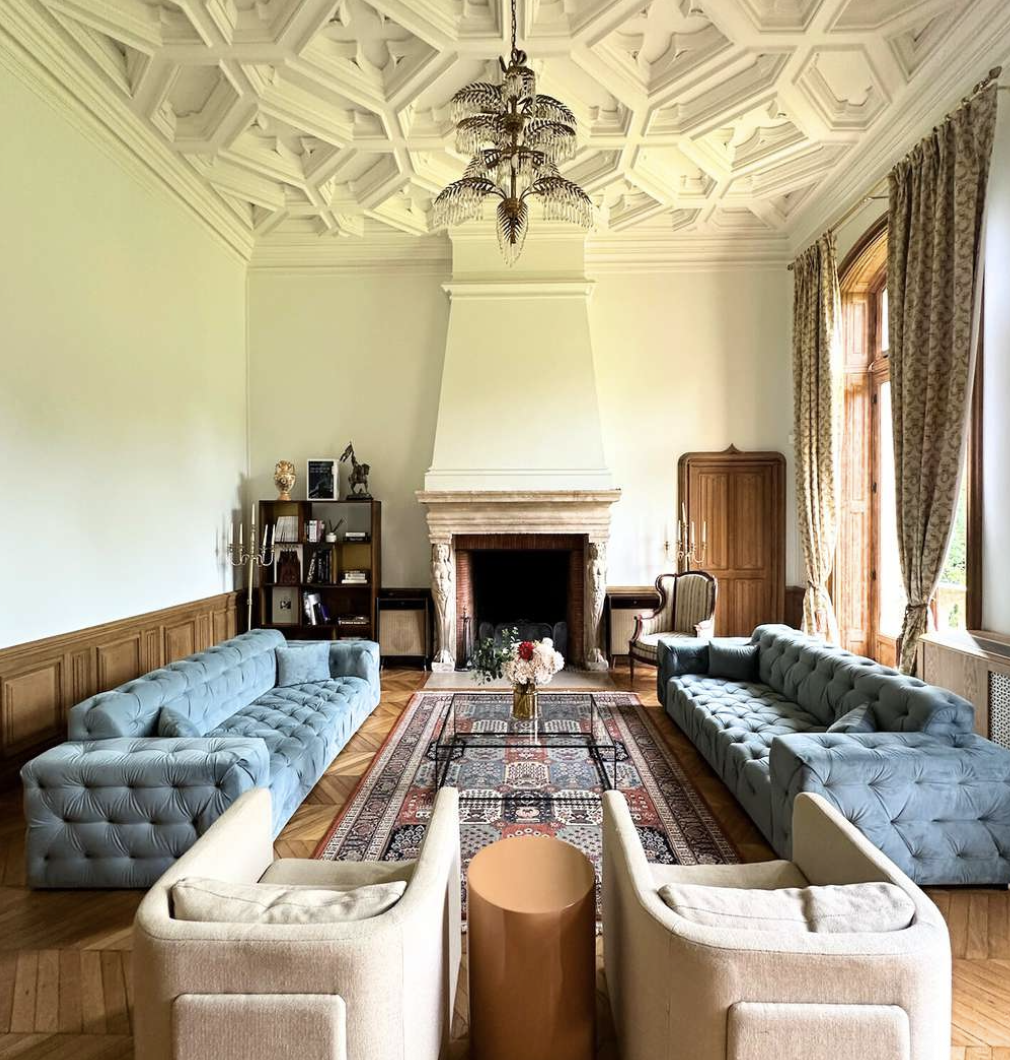 Elegant living room with two large, tufted, blue sofas facing each other, a white armchair in front, a dark wooden bookcase on the left with decor, a wooden cabinet on the right, a fireplace in the center with floral arrangements on top, a colorful p