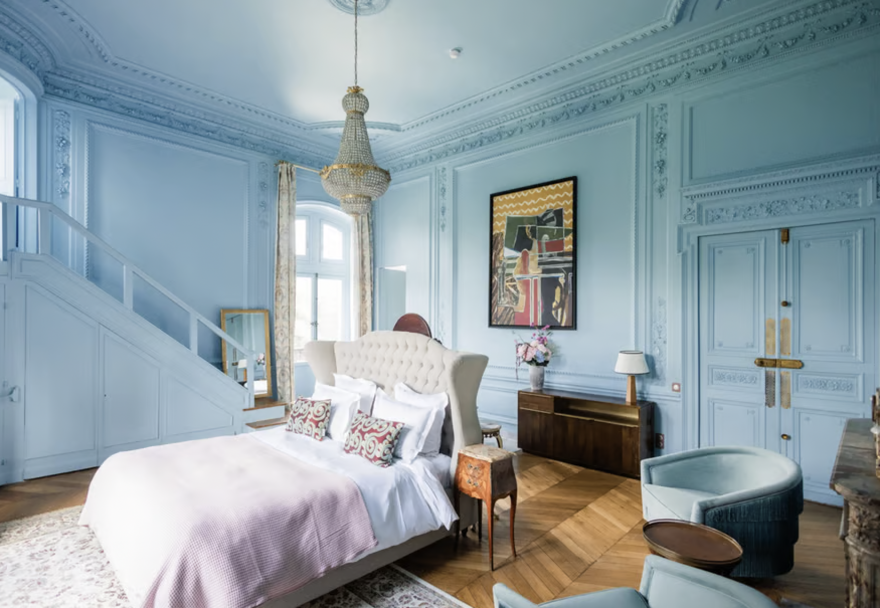 A luxurious bedroom with blue paneled walls and elaborate crown molding, featuring a chandelier, a tufted headboard bed with white and patterned pillows, a wooden nightstand with a lamp and flowers, artworks, and seating areas with pastel-colored cha