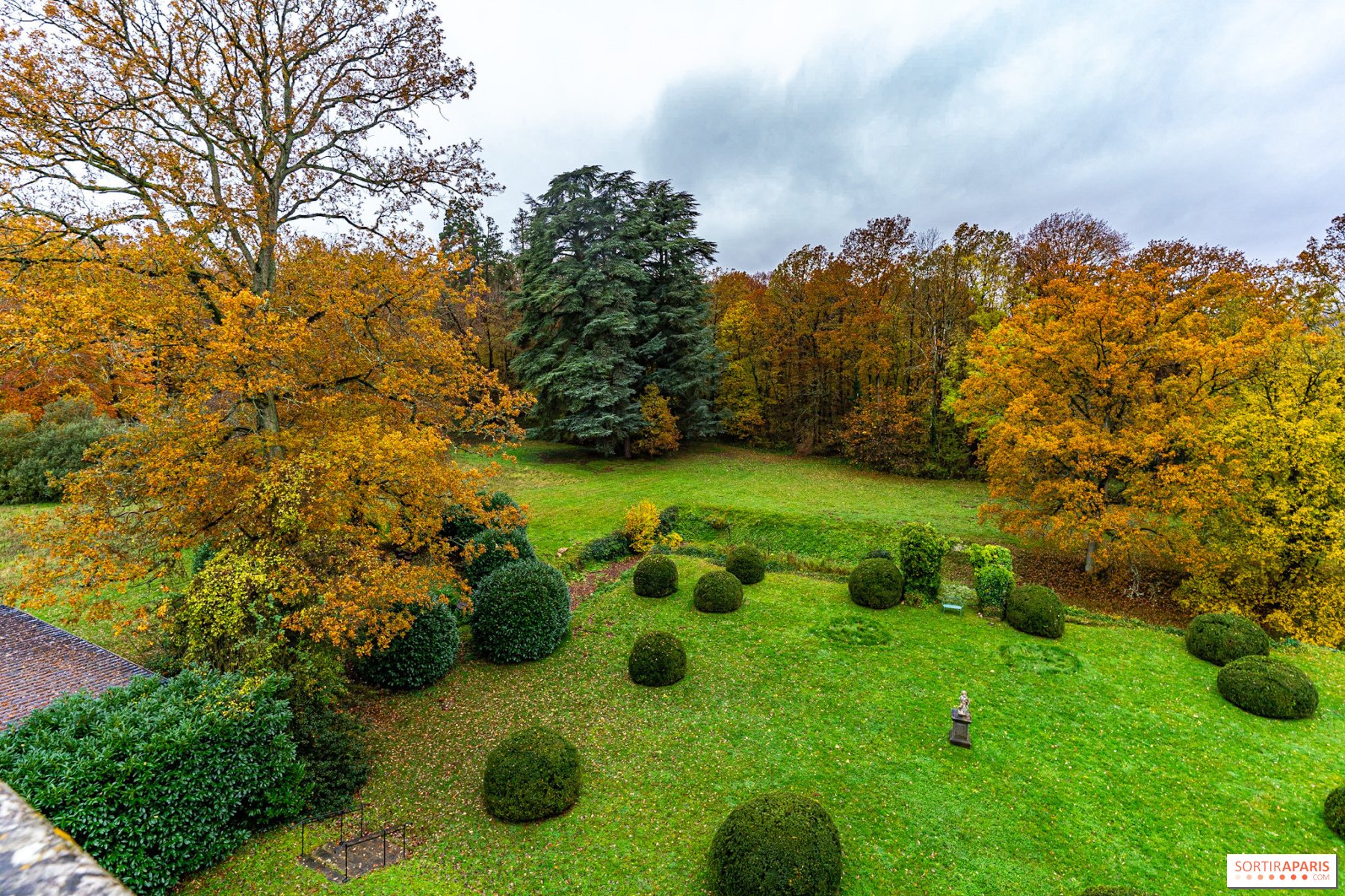 A lush green garden with round bushes, a variety of large trees with fall foliage, and a cloudy sky overhead.