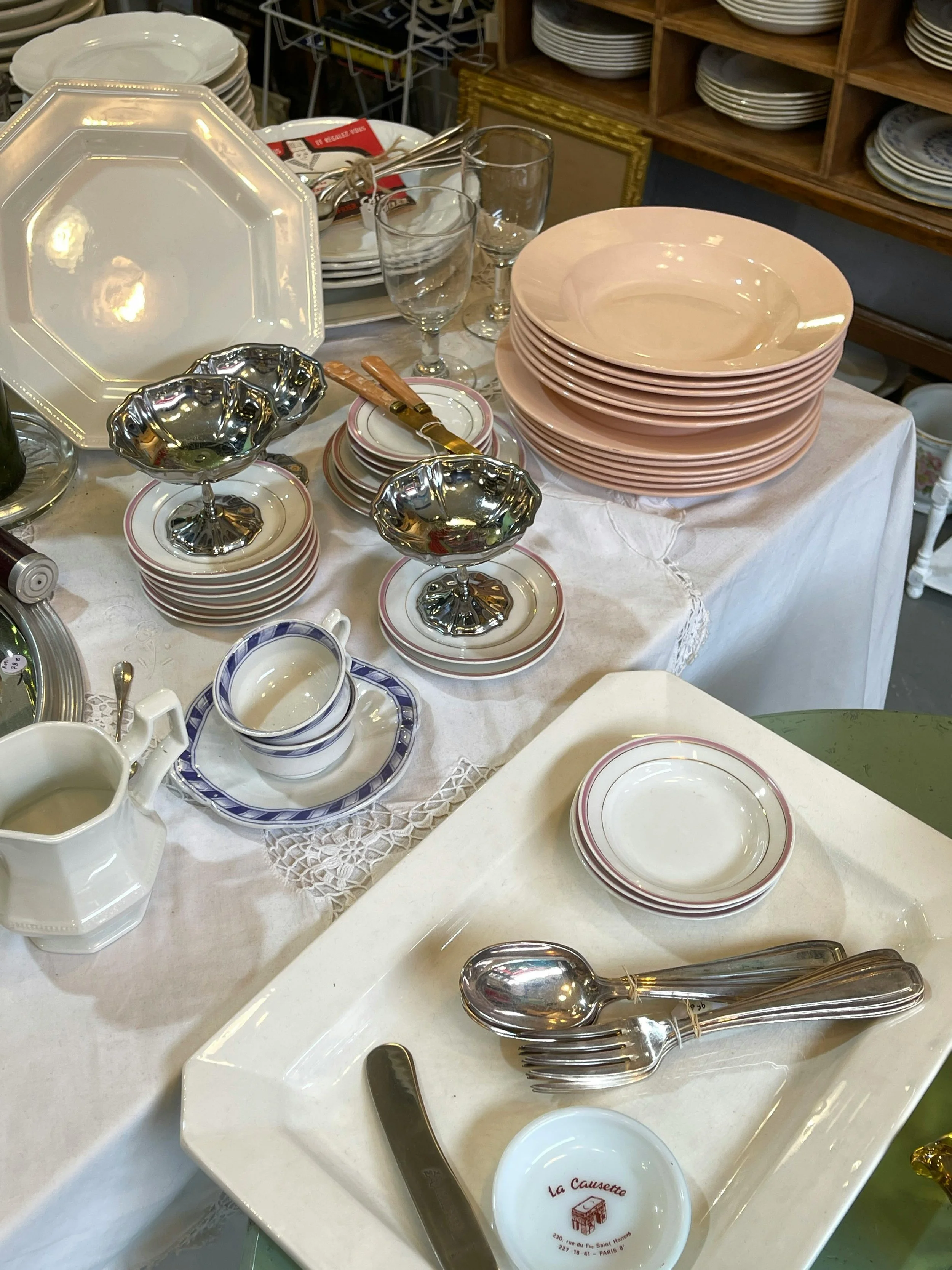 Display of various dishes and utensils on a table, including plates, bowls, silverware, and glasses, with shelves of plates in the background.