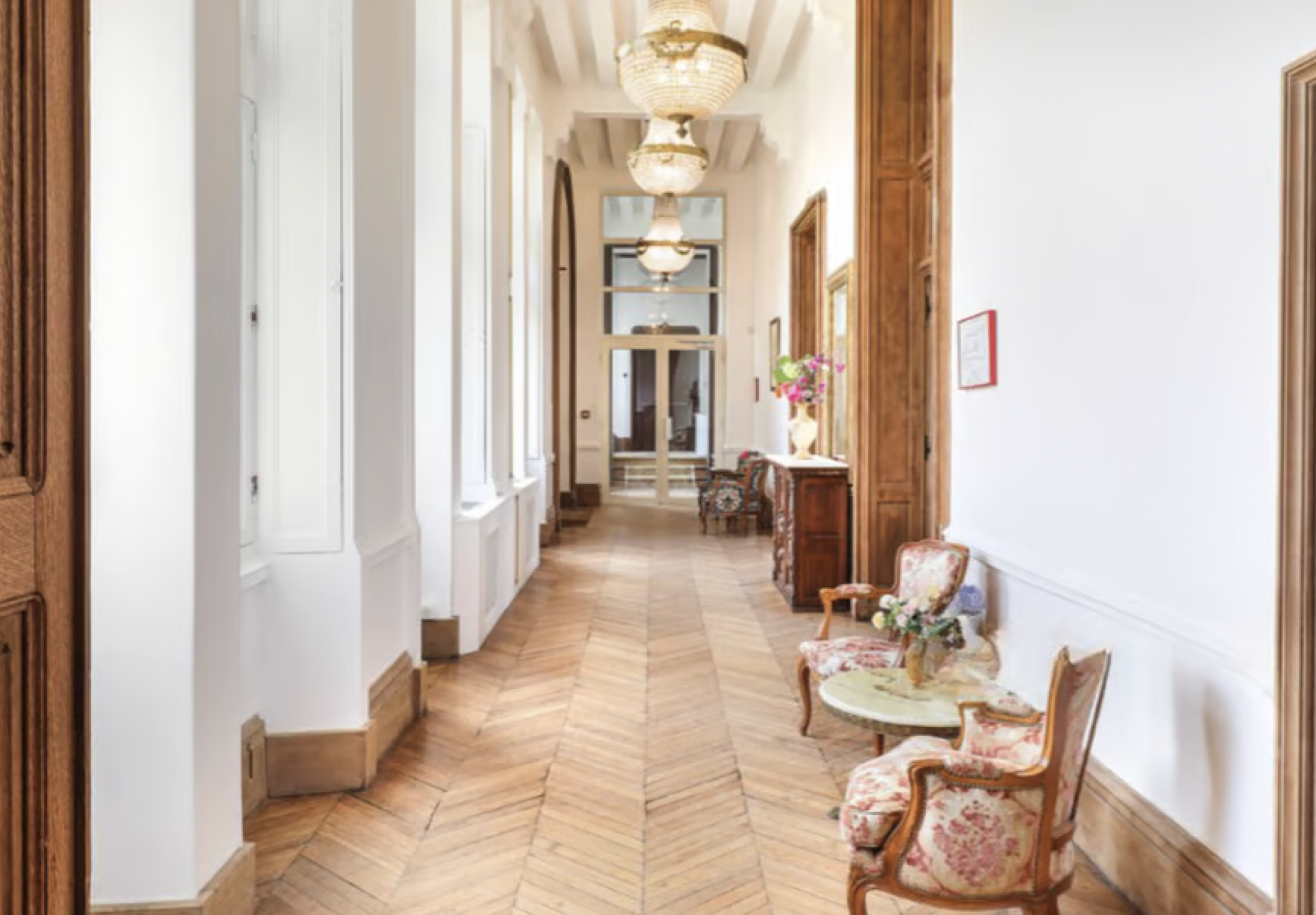 Elegant hallway with wooden flooring, white walls, large windows, and vintage chairs with floral upholstery along the right wall. A chandelier hangs from the ceiling, and there are decorative elements like a vase of flowers and framed artwork.