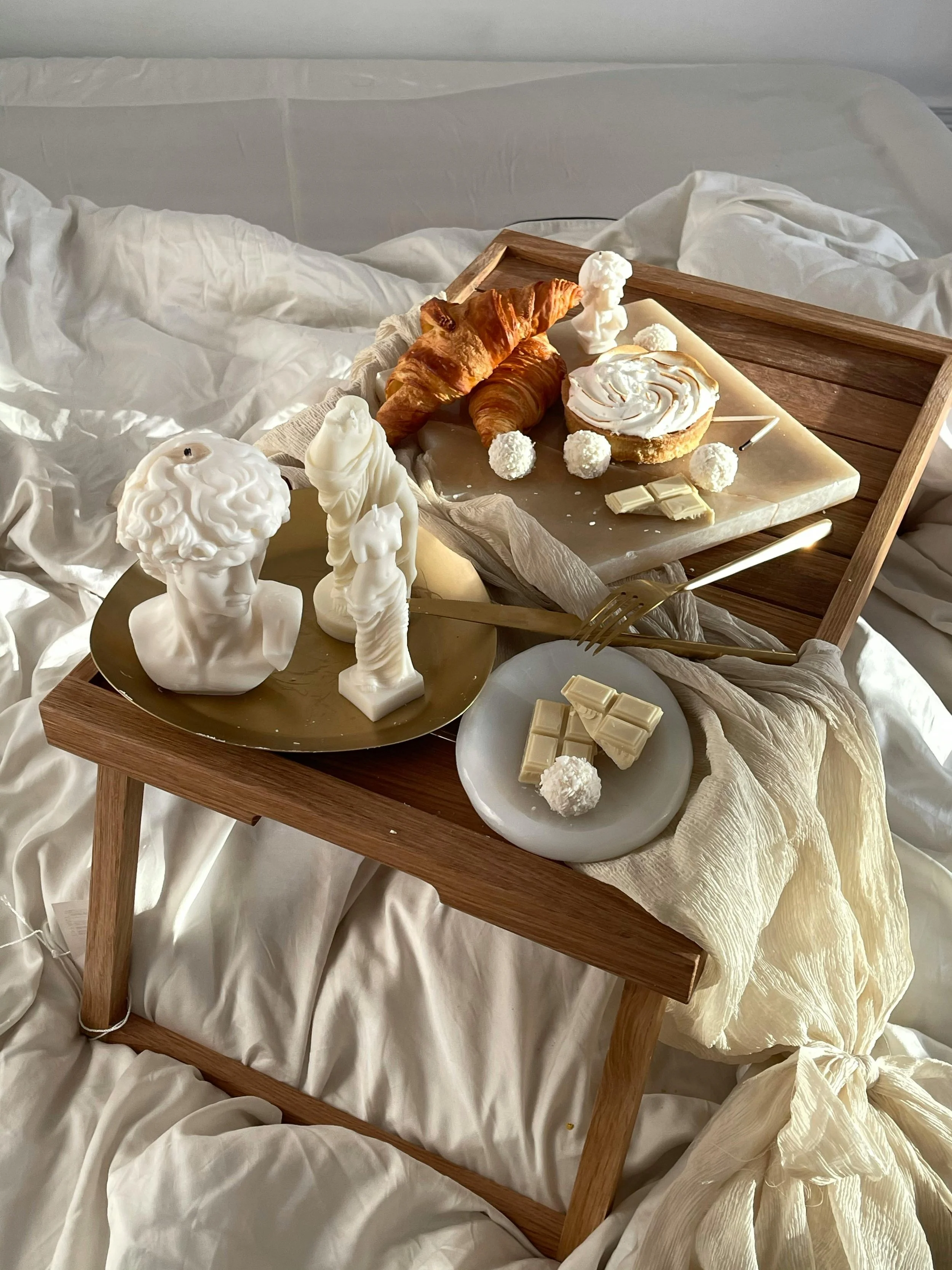 A wooden tray on a bed with white bedding features croissants, white chocolate, a small bust sculpture, white truffles, a tart with whipped cream, and white chocolate pieces, accompanied by decorative white bust statues nearby.