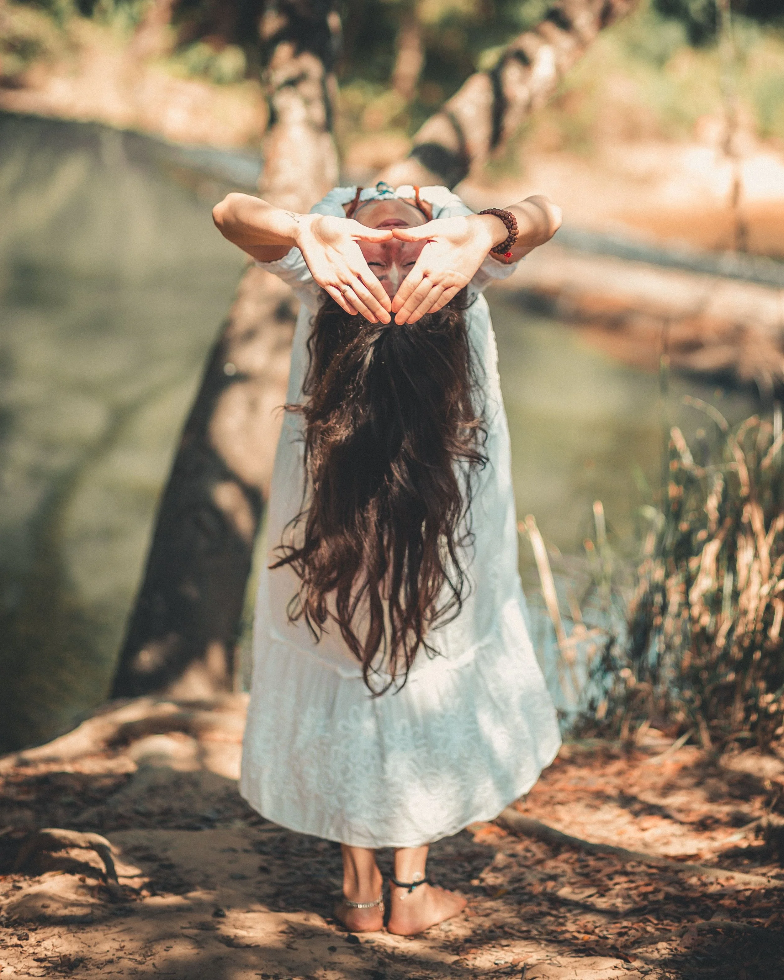 A person with long hair wearing a white lace dress standing barefoot on leaf-covered ground, making a heart shape with hands over their face, with trees and sunlight in the background.