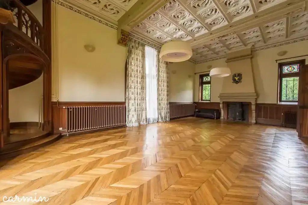 An empty, spacious living room with a parquet wooden floor, ornate ceiling, large window with floral curtains, fireplace with a decorative crest on the wall above, and two pendant lights hanging from the ceiling.