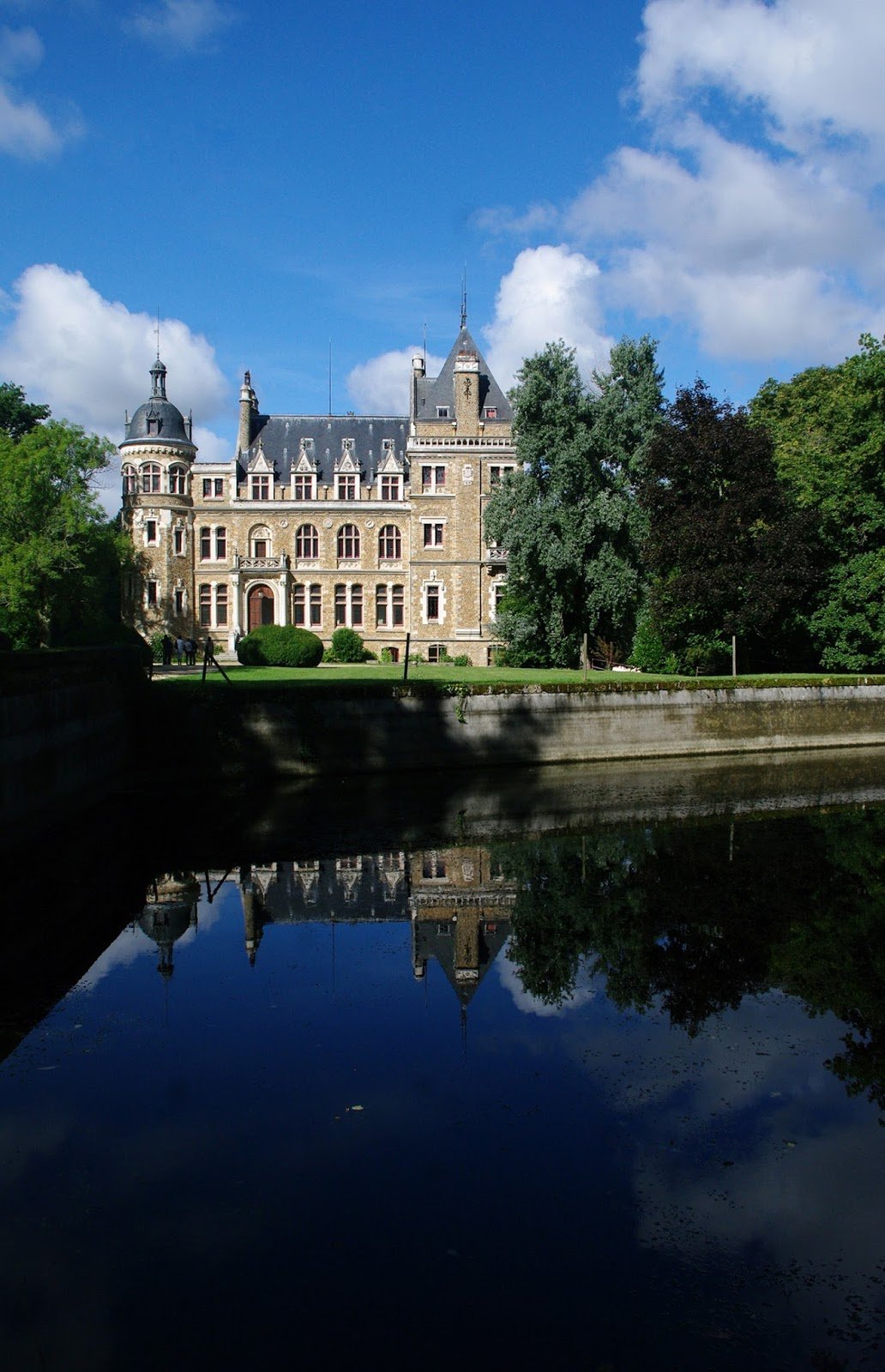 A large castle with turrets and a slate roof, surrounded by trees and a green lawn, with a body of water in the foreground reflecting the castle and sky.