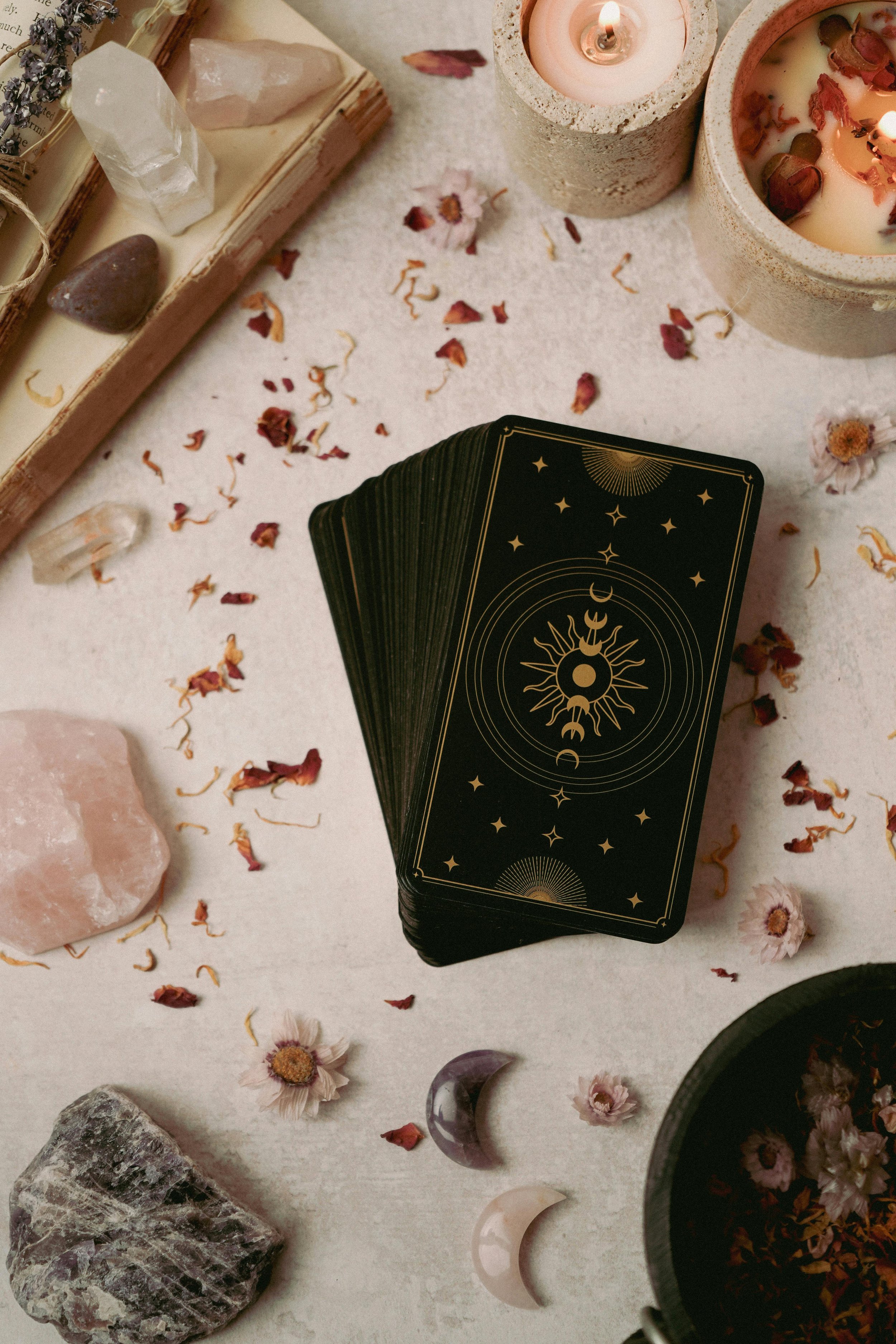 A deck of tarot cards with celestial designs, surrounded by candles, dried flowers, crystals, and stones on a light surface, with scattered dried flower petals.
