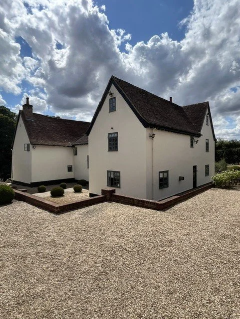 Driveway-side view of a fully restored farmhouse exterior in Dedham, Essex, featuring Bedec Gardenia masonry paint with contrasting black facias and soffits and traditional green windows.