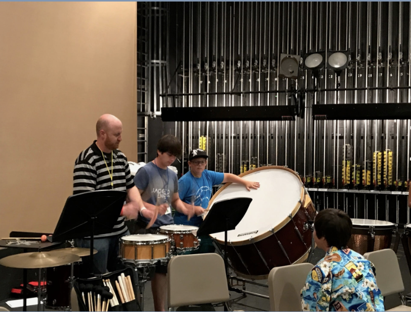 Group of children and an adult playing drums in a music room with percussion instruments and a black metal backdrop.