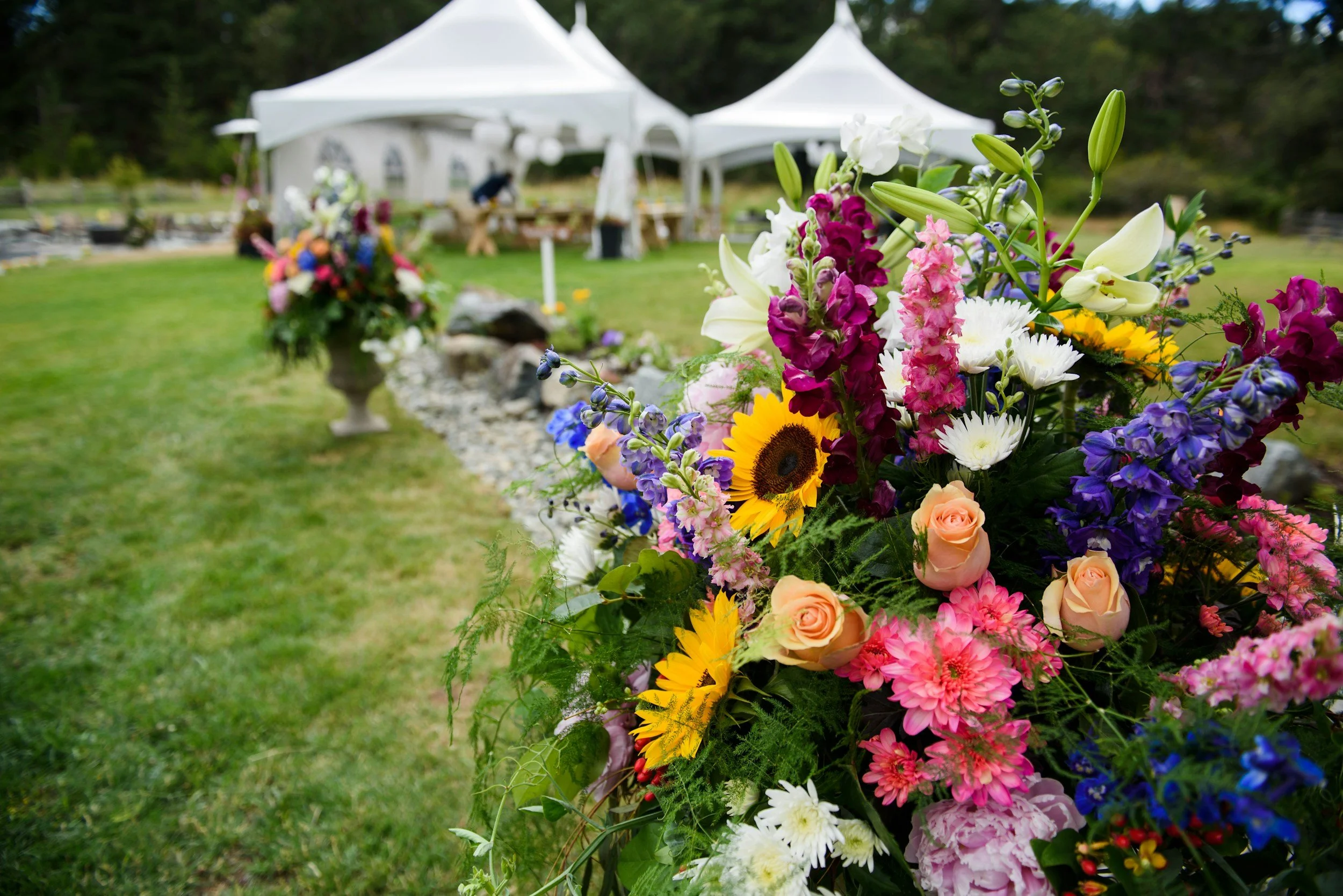 A large, colorful flower arrangement featuring sunflowers, roses, lilies, and other blooms in front of white tents at an outdoor wedding.