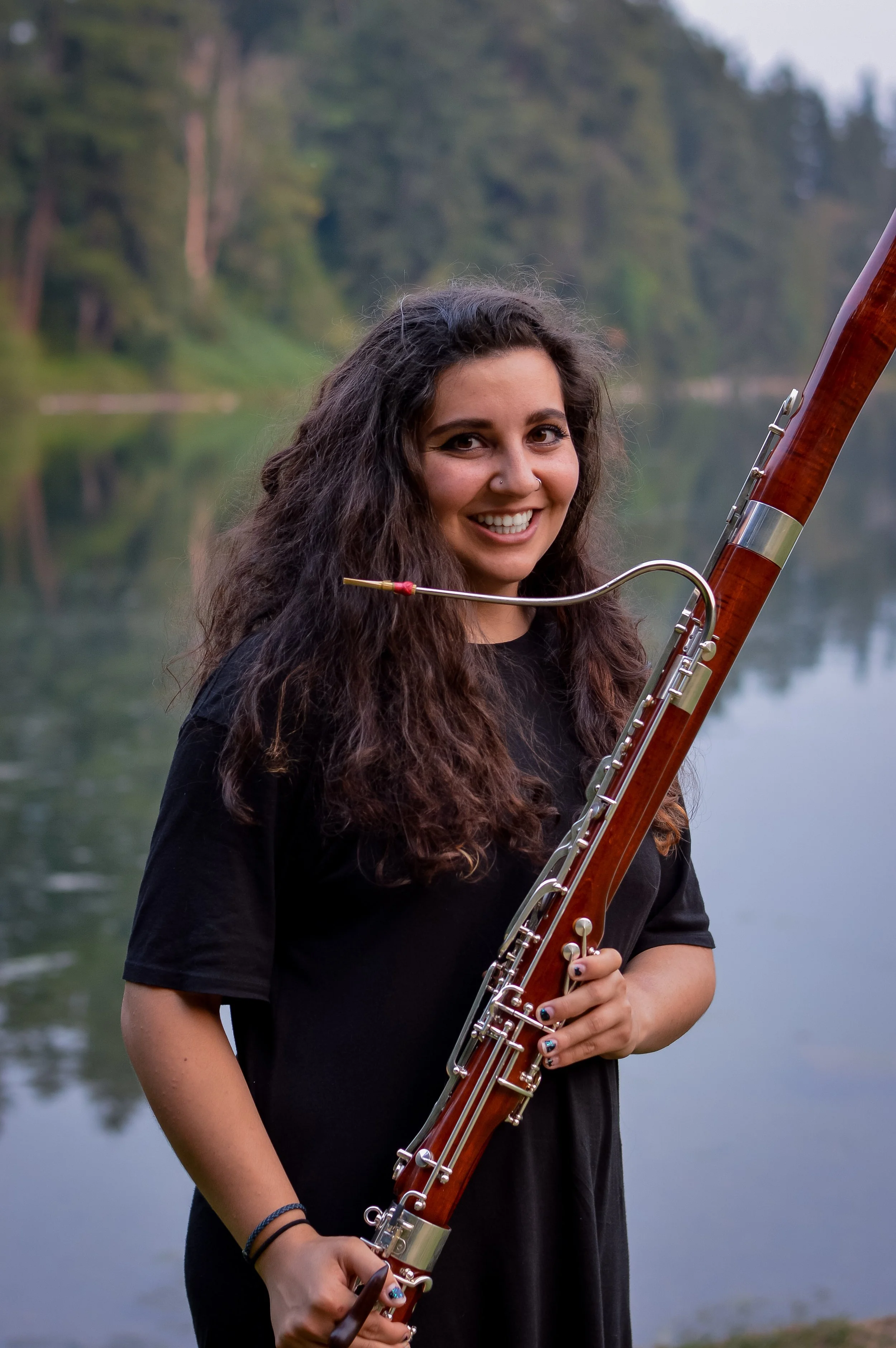 A young woman with long, curly dark hair smiling and holding a bassoon, standing in front of a calm body of water surrounded by green trees.