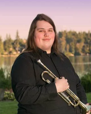 Person holding a trumpet outdoors with trees and water in the background.