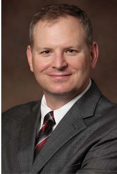 A man in a gray suit with a white shirt and a red striped tie, smiling at the camera with a brown background.