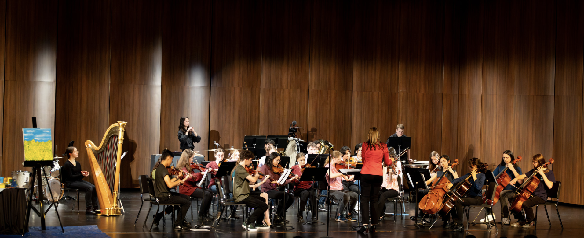 Orchestra performing in a concert hall with wooden walls, featuring musicians playing violins, cellos, and other string instruments, led by a conductor in a red jacket.