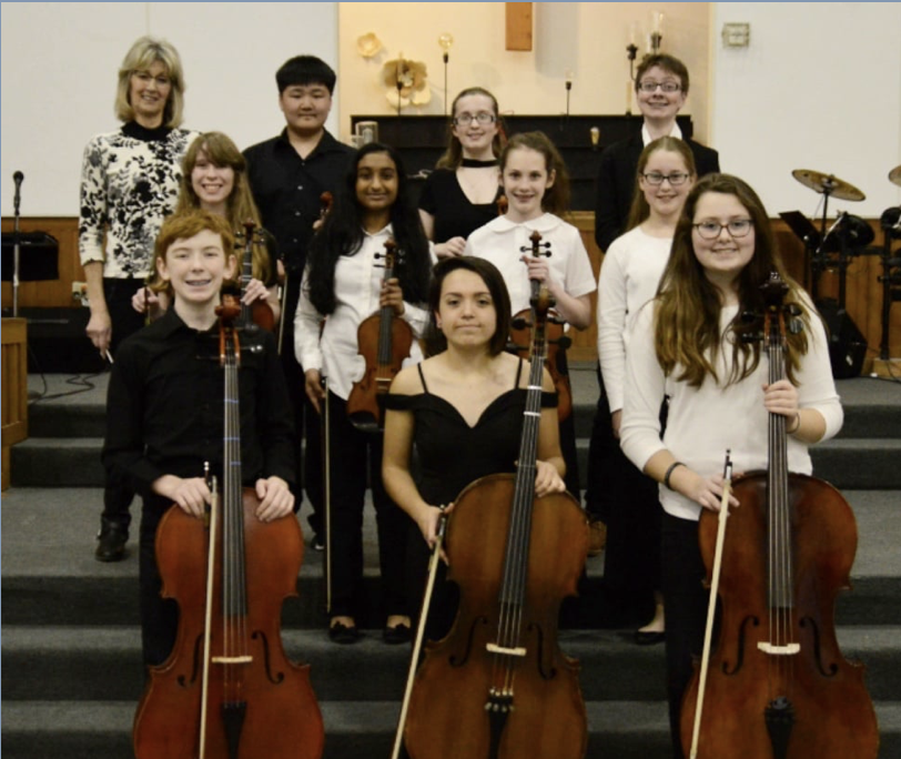 Group of young students with string instruments, including cellos and violins, standing on a stage with a teacher in the background after a musical performance.