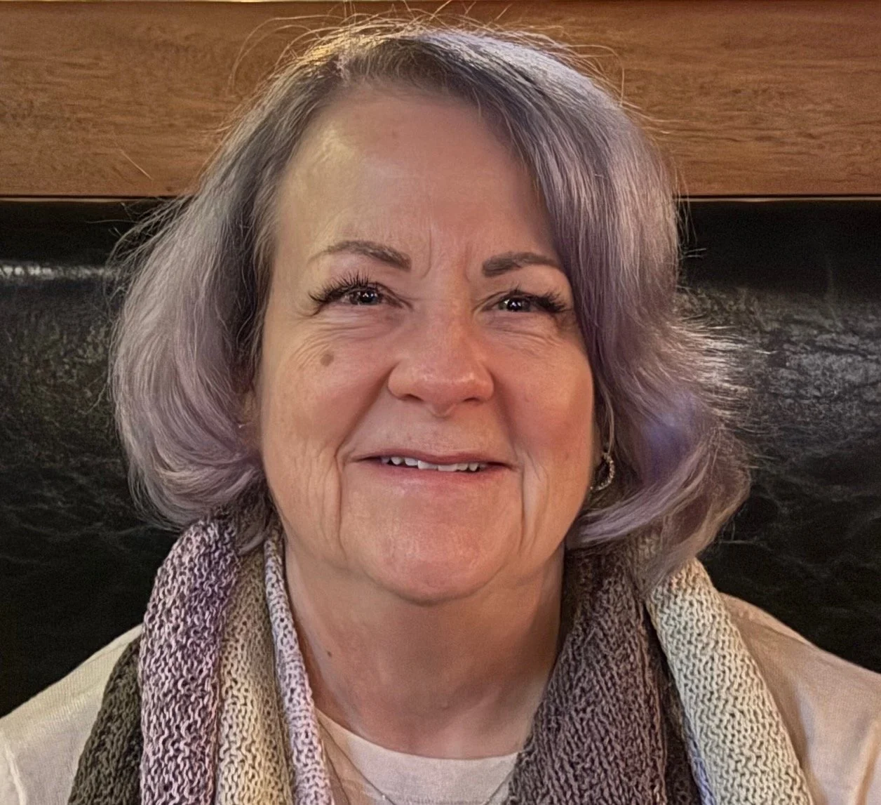 A smiling middle-aged woman with short, wavy gray hair, wearing a multicolored scarf and earrings, sitting in front of a wooden and black background.
