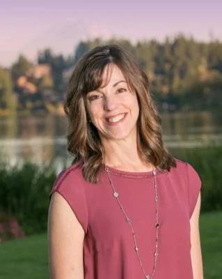 A smiling woman with shoulder-length brown hair wearing a pink sleeveless top and a necklace, standing outdoors near a body of water with greenery and trees in the background during sunset.