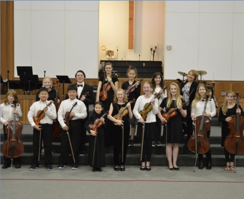 Youth orchestra members posing with their string instruments on stage in a church or school auditorium.