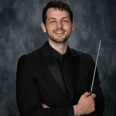 A man in a black suit holding a conductor's baton, standing in front of a dark, textured background.