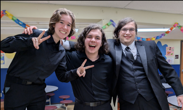 Three young men in formal attire posing happily in an indoor decorated room, making peace signs and smiling
