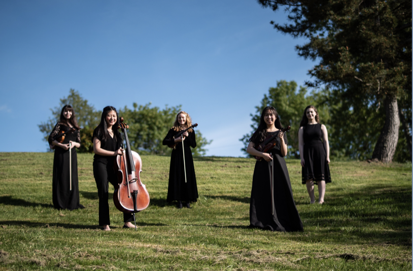 Five women in black dresses standing on a grassy hill, four holding musical instruments, outdoors on a sunny day with trees and blue sky in the background.