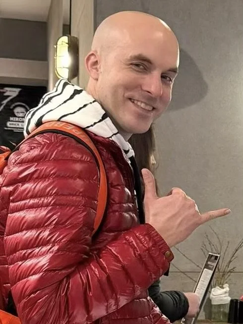 A smiling man with a bald head wearing a red puffer jacket and striped hoodie, giving a shaka sign with his right hand.