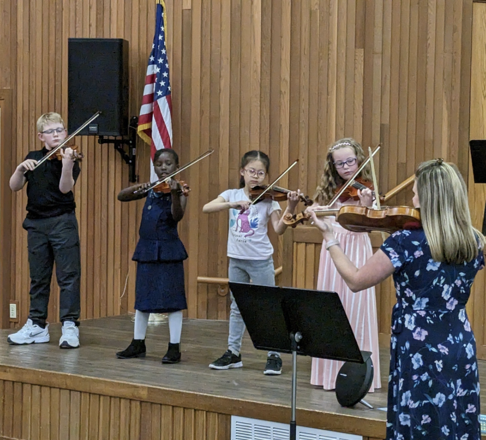 Five children playing violins on a wooden stage, with a woman conducting, in a room with wood-paneled walls and an American flag in the background.