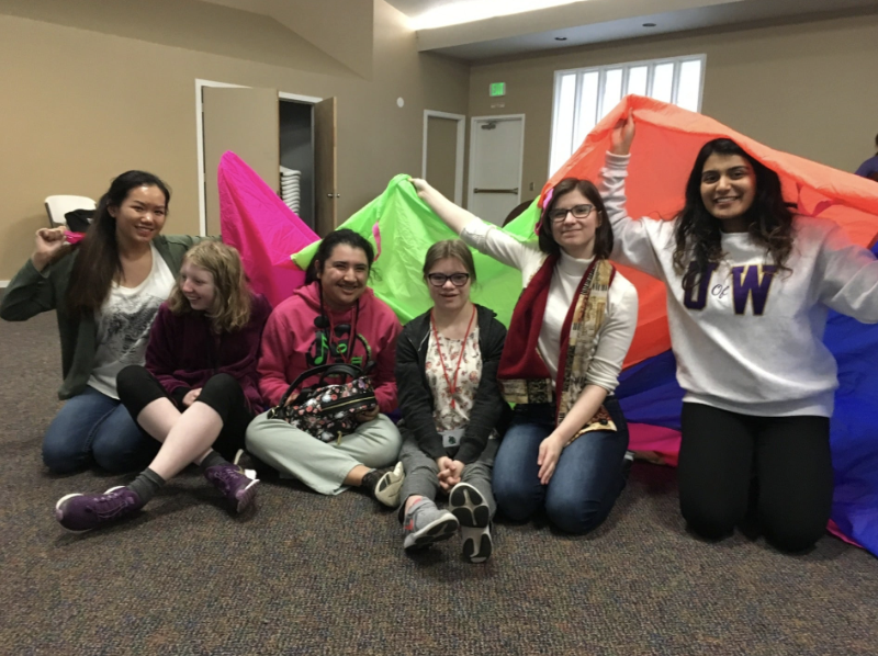 Group of six smiling young women sitting on the floor with colorful parachute in a room.