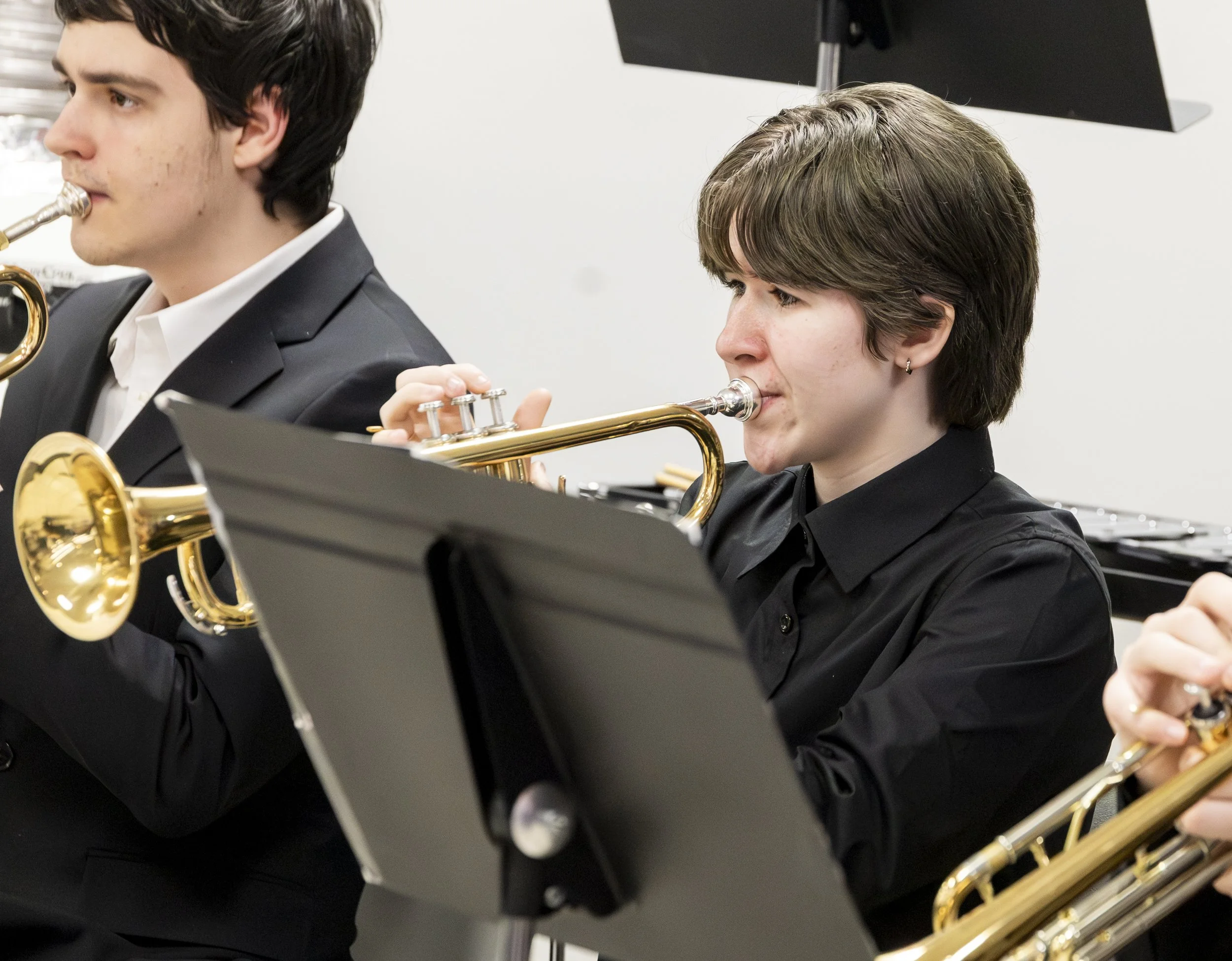 Two young students in black formal suits playing instruments during a music performance, with music stands in front of them, in an indoor setting.
