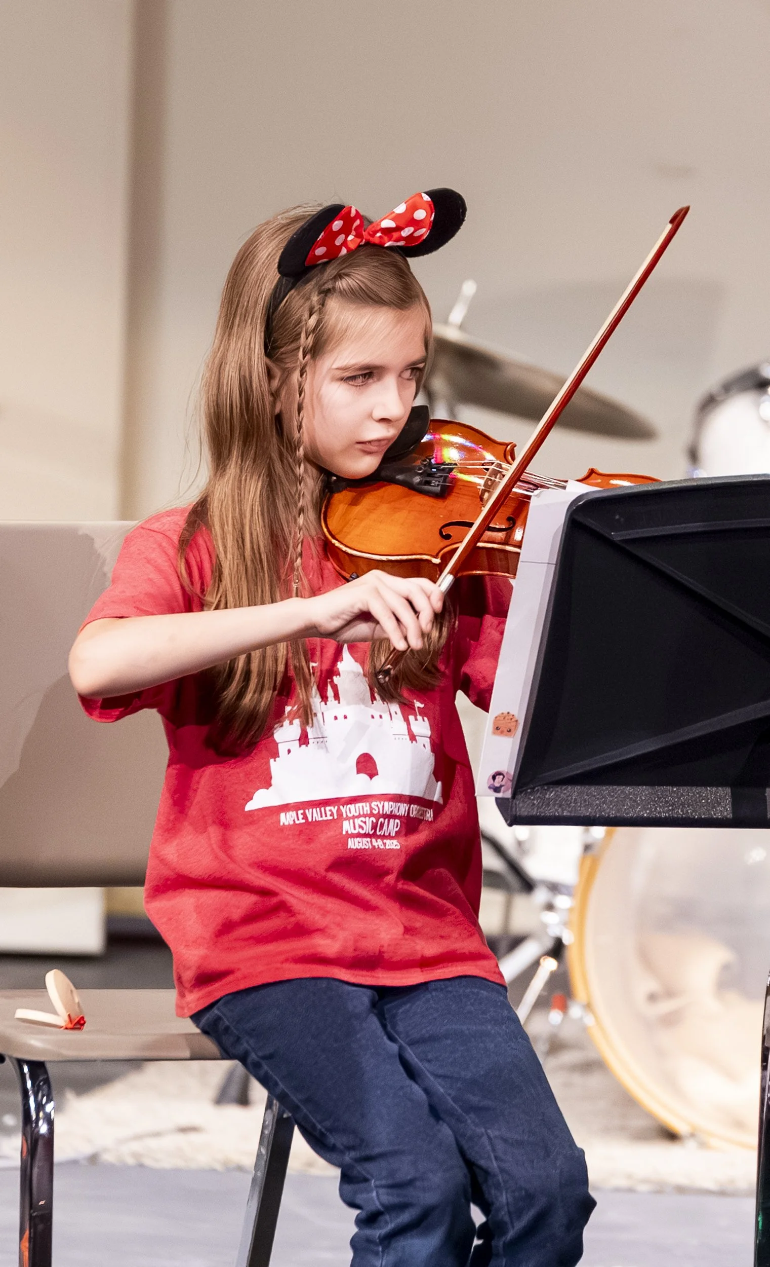 A young girl with long blonde hair wearing a Minnie Mouse headband playing the violin while seated, with a music stand in front of her.