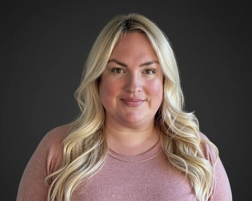 A woman with long blonde hair and a pink shirt, smiling against a dark gray background.