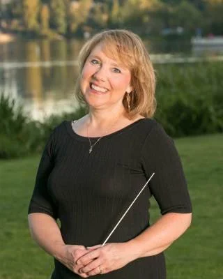 Woman smiling outdoors by a lake holding a conductor's baton