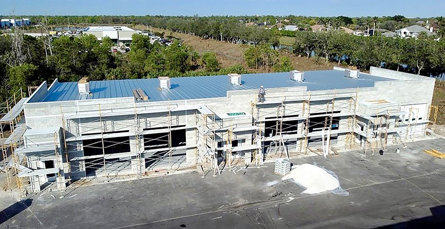 Construction site of a large, single-story building with scaffolding, a metal roof, and unfinished concrete walls, situated on an asphalt lot with a pile of white material nearby. Trees and other buildings are visible in the background.