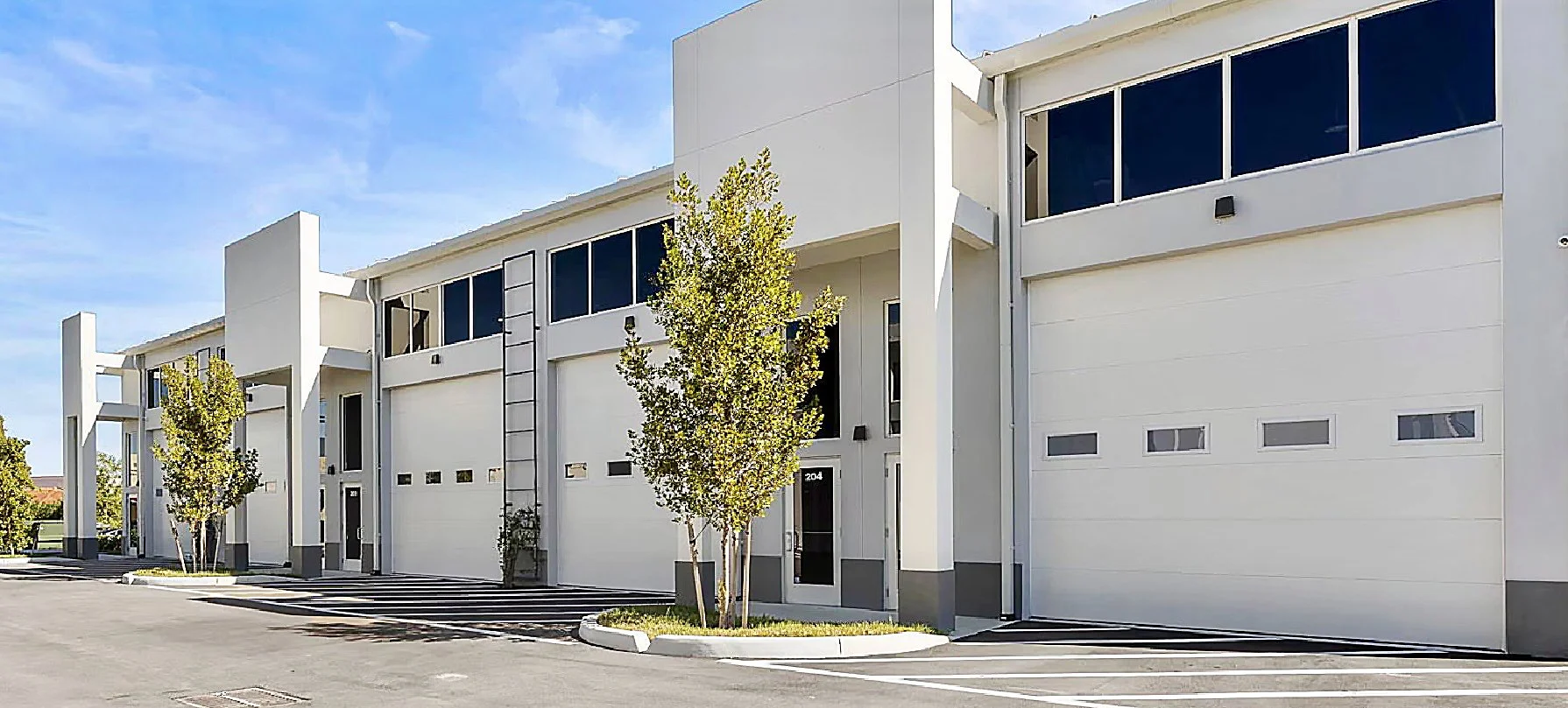 Modern white townhouse apartments with large garage doors, trees, and clear blue sky.