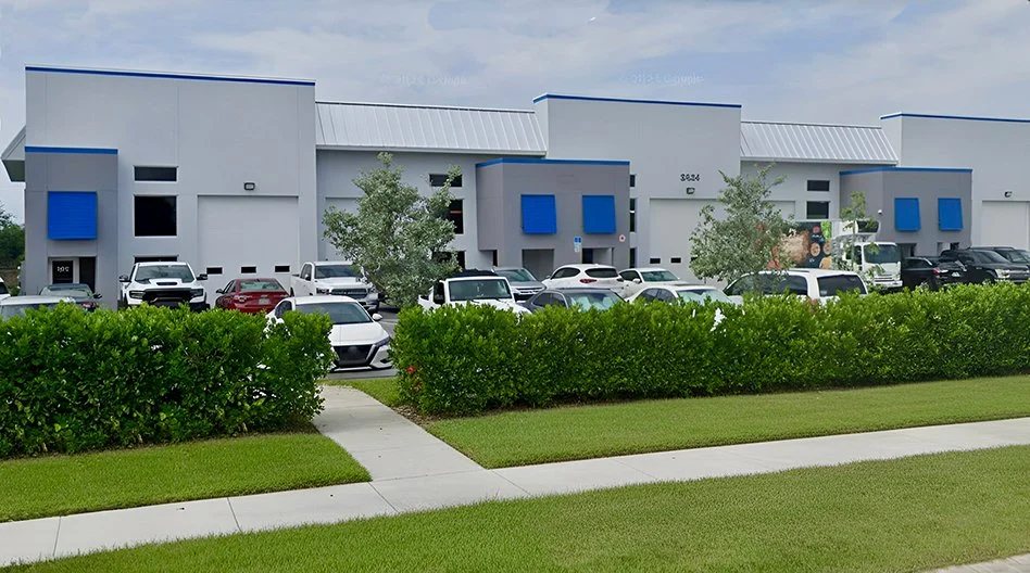 Exterior view of a commercial building with parking lot, several cars, and bushes in front.