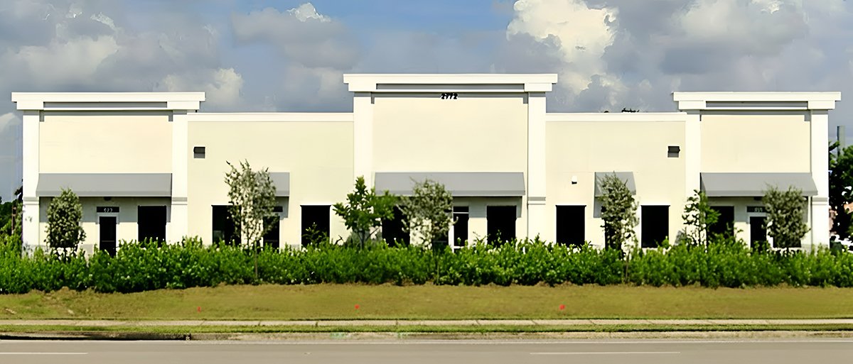 A modern white commercial building with multiple storefronts and gray awnings, surrounded by green bushes and trees under a partly cloudy sky.