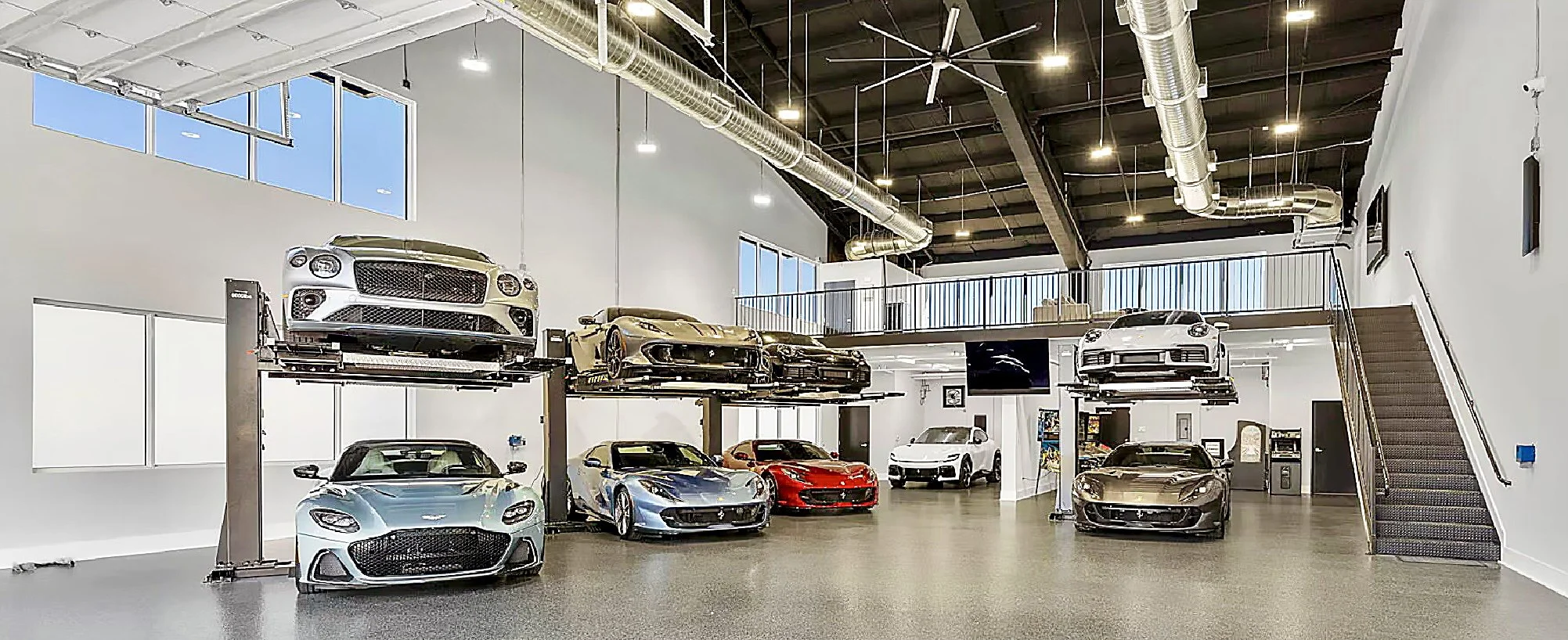Luxury cars displayed on elevated and ground-level car lifts inside a modern showroom with high ceilings, large windows, and industrial-style ductwork.