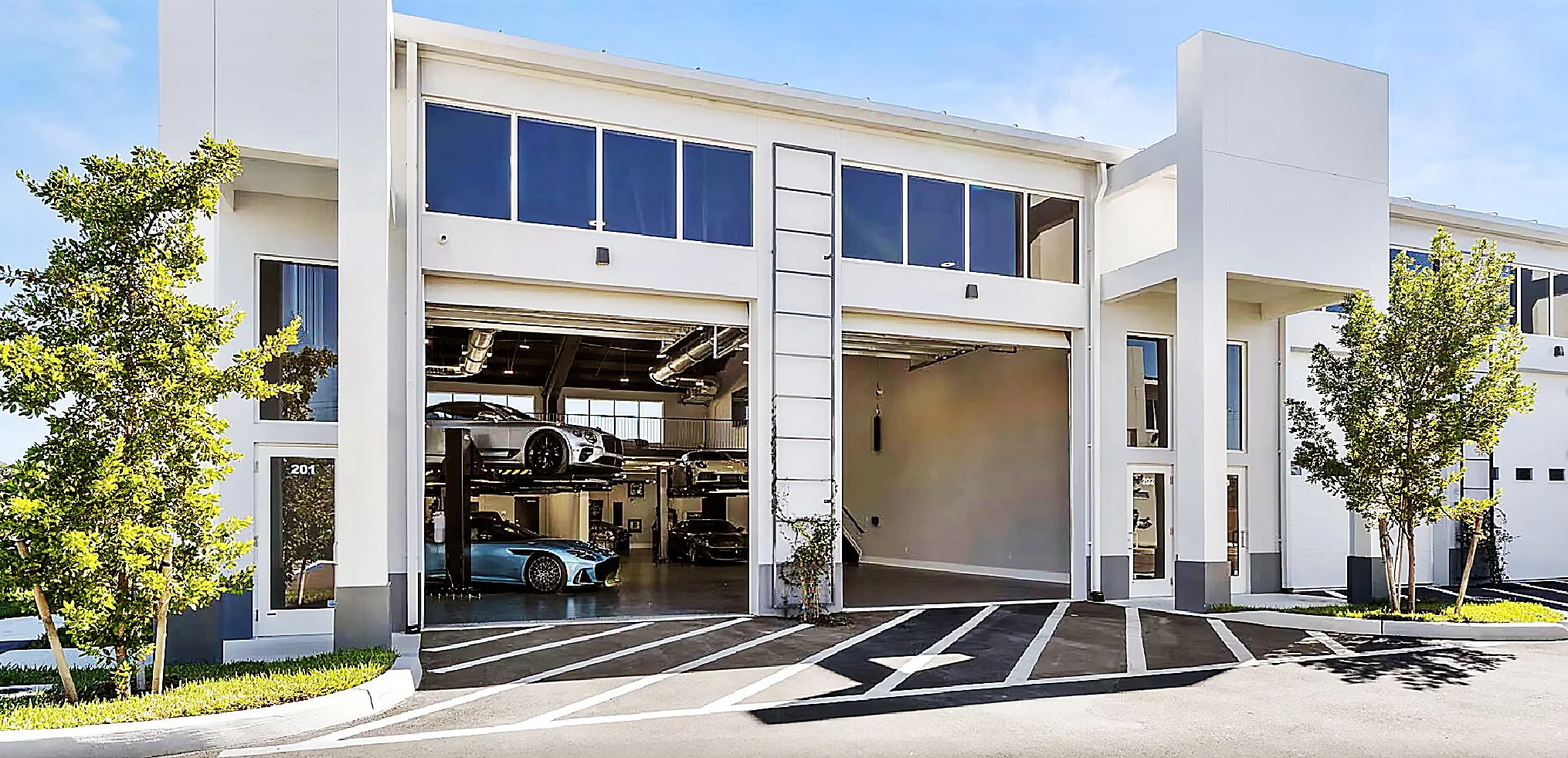 Modern two-story building with an automotive garage, two cars parked inside, and trees in the front yard.
