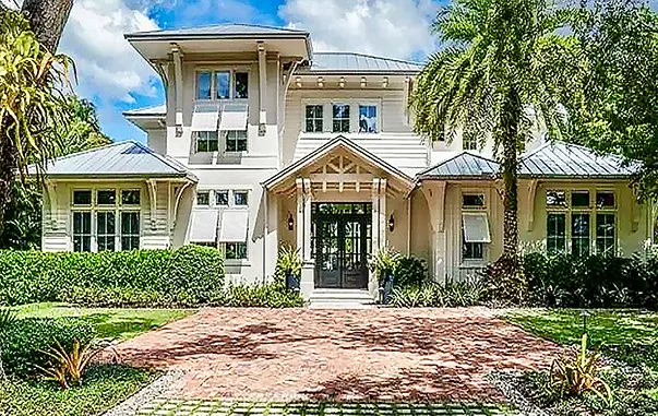 Large white house with multiple stories, surrounded by palm trees, shrubs, and a brick driveway.