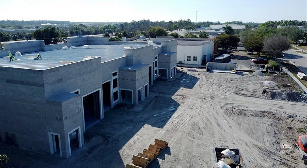 Construction site of a large building with a flat roof, surrounded by dirt and construction materials, in a suburban area with trees and parked cars.