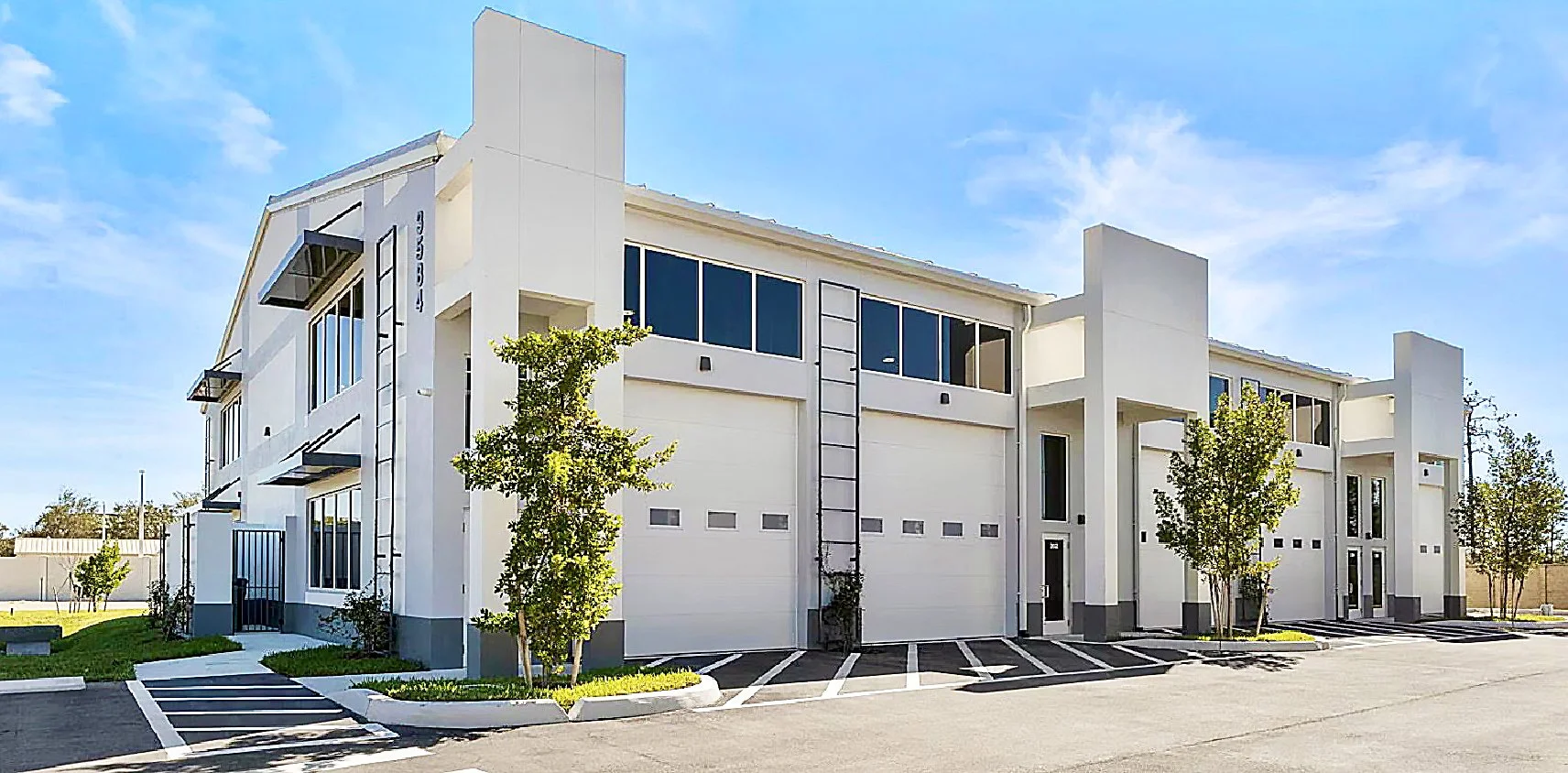 Modern white multi-unit residential building with garage doors, parking spaces, and small trees in front, under a clear blue sky.