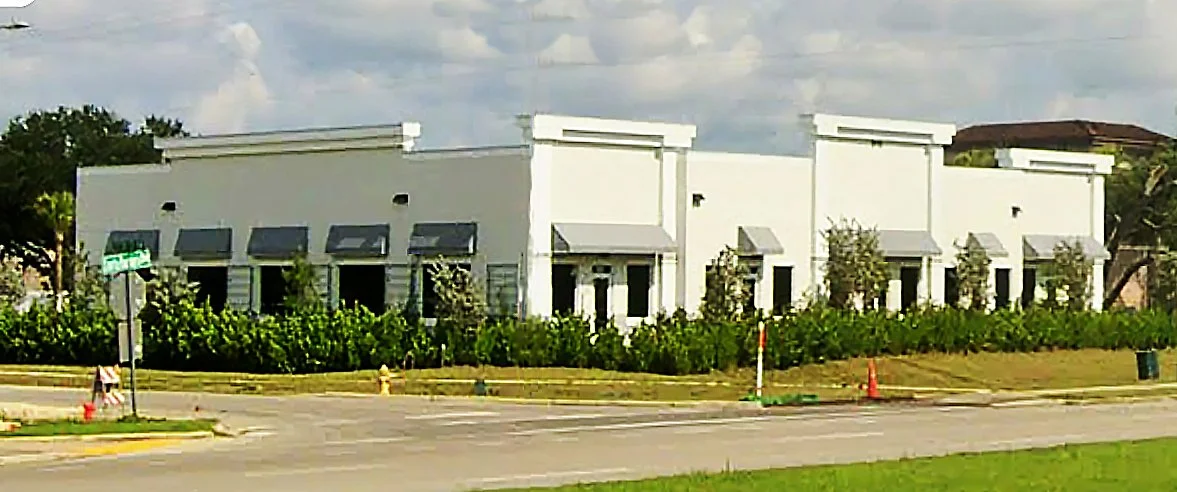 A white commercial building with multiple windows and awnings, surrounded by greenery and located on a street corner.